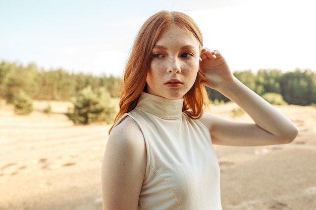 Portrait of young woman with freckles touching long wavy ginger\
hair and looking away