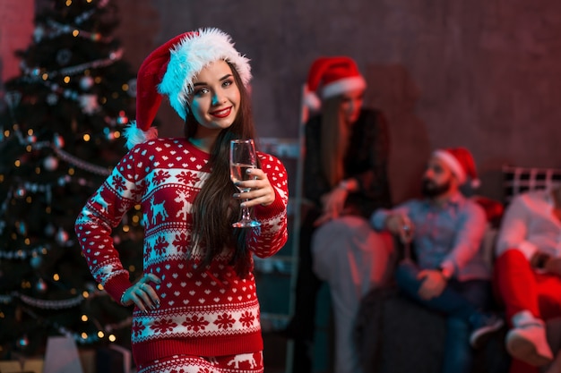 Portrait of a young woman with a glass of champagne at home on the foreground. Beautiful brunette in a Santa hat, red costume with deers