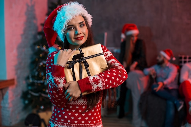 Portrait of a young woman with a gold giftbox at home on the foreground. Beautiful brunette in a Santa hat, red costume with deers