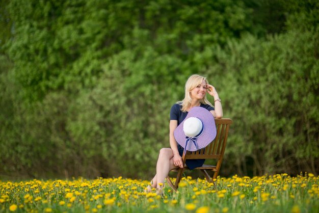 Portrait of young woman with hat sitting on chair at dandelion meadow