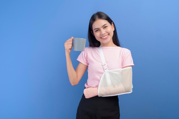 A portrait of young woman with an injured arm in a sling having a cup of coffee over blue background in studio, insurance and healthcare concept