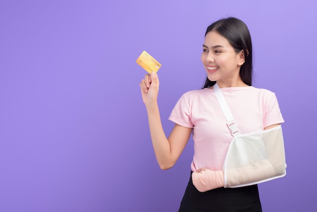 Portrait of young woman with an injured arm in a sling holding a credit card over purple wall