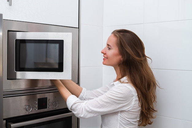 Photo portrait of a young woman with long hair cooking in a microwave oven opening the door and taking out food