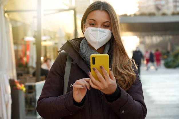 Ritratto di una giovane donna con maschera medica che cammina in città utilizzando lo smartphone al tramonto