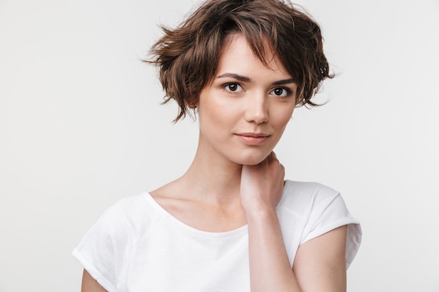 Portrait of young woman with short brown hair in basic t-shirt  while standing isolated over white wall