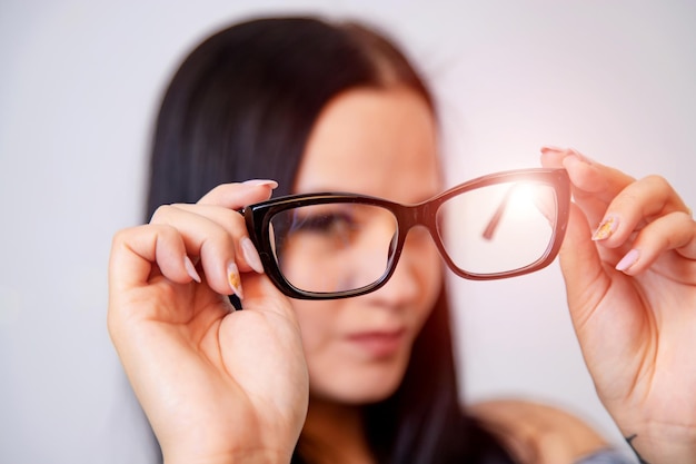 Портрет молодой женщины с очками в руках Размытый белый фон Девушка смотрит в очки Длинноволосая брюнетка красивая девушка и очки в черной оправе Крупным планом