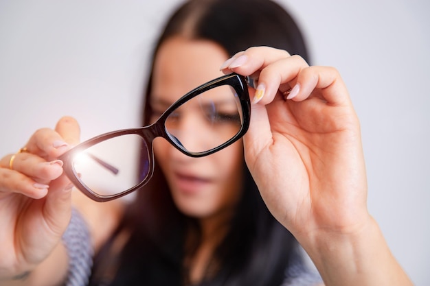 Portrait of a young woman with spectacles in hands Blurred white background Girl looks through eyeglasses Longhaired brunette beautiful girl and black framed eyeglasses Closeup