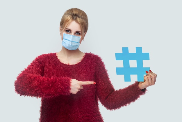 Portrait of young woman with surgical medical mask in red blouse standing big blue hashtag sign and pointing finger, social media concept. Indoor, isolated, studio shot, copy space, gray background