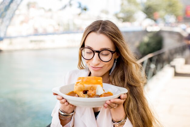 Portrait of a young woman with traditional portuguese sandwich with meat called francesinha on the landscape background with bridge in Porto city, Portugal