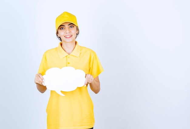 Portrait of a young woman in yellow uniform holding an empty white speech bubble cloud . 