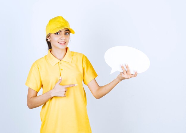 Portrait of a young woman in yellow uniform pointing at an empty speech bubble . 