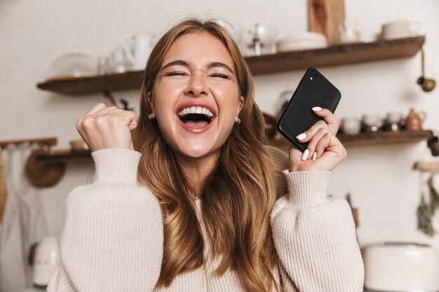 portraitcloseup of excited caucasian woman making winner gesture and using cellphone in cozy kitchen