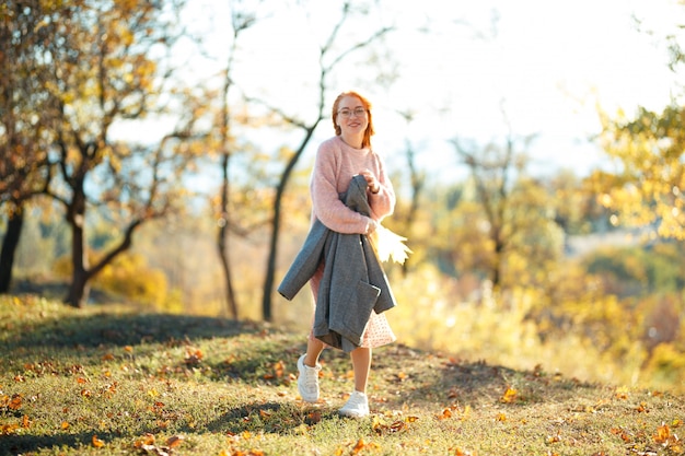 Ritratti di un'affascinante ragazza dai capelli rossi con un viso carino. ragazza che posa nel parco di autunno in un maglione e una gonna color corallo. la ragazza ha un umore meraviglioso