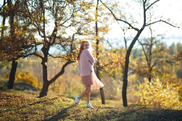 Portraits of a charming red-haired girl with a cute face. Girl posing in autumn park in a sweater and a coral-colored skirt. In the hands of a girl a yellow leaf