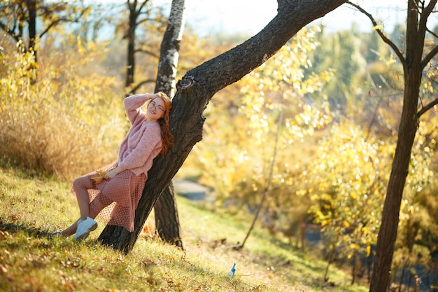 Portraits of a charming red-haired girl with a cute face. Girl posing in autumn park in a sweater and a coral-colored skirt. In the hands of a girl a yellow leaf