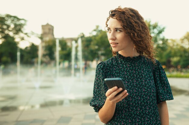 Portraits of a charming red-haired girl with a pretty face. The girl holds a mobile phone in her hands and looks to the side. She has a great mood and a sweet smile