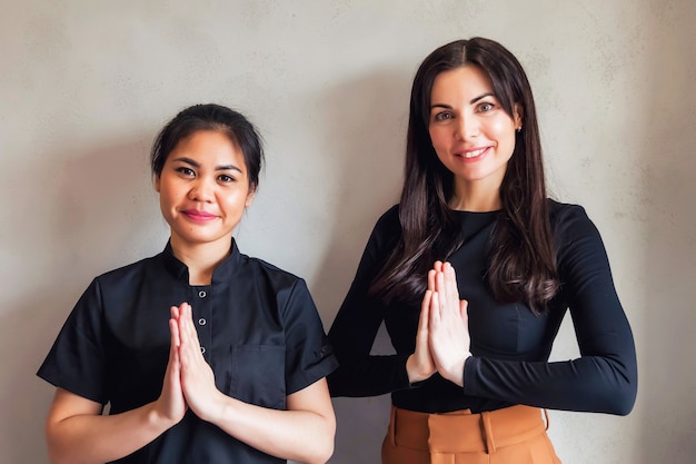 Portraits of females administrator and master masseur with greeting  standing at grey wall background. Young woman manager and asian Bolivian professional worker dressed uniform in modern SPA salon