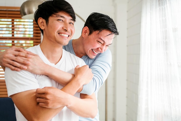 Portraits of happy millennial Asian gay couple hugging and embracing smiling and laughing in a white curtain window in the living room at LGBT multirelationship home An enjoy gay couple concept