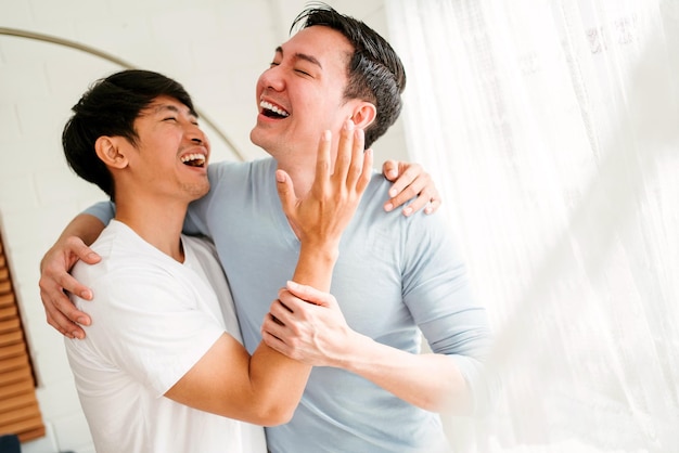 Portraits of happy millennial Asian gay couple hugging and embracing smiling and laughing in a white curtain window in the living room at LGBT multirelationship home An enjoy gay couple concept