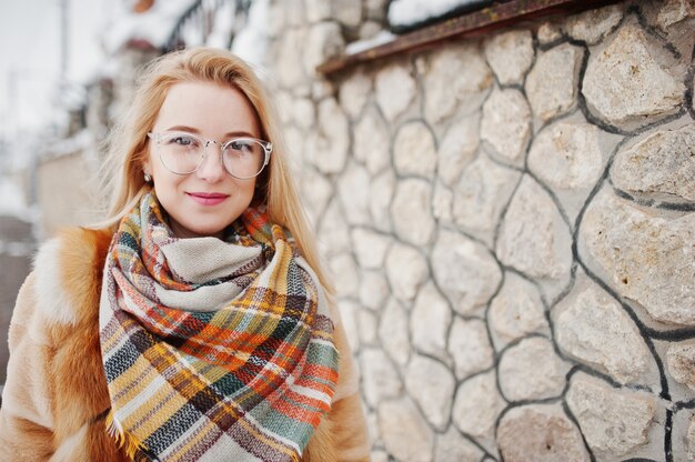 Portraiy of blonde girl in glasses, red fur coat and scarf at winter day.