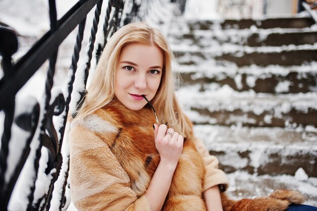 Portraiy of blonde girl in glasses, red fur coat and scarf at winter day.