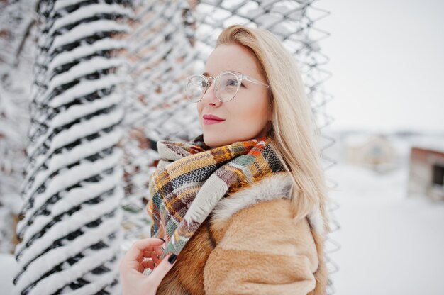 Portraiy van blondemeisje in glazen, rode bontjas en sjaal bij de winterdag.