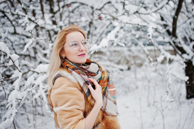 Portraiy van blondemeisje in glazen, rode bontjas en sjaal bij de winterdag.