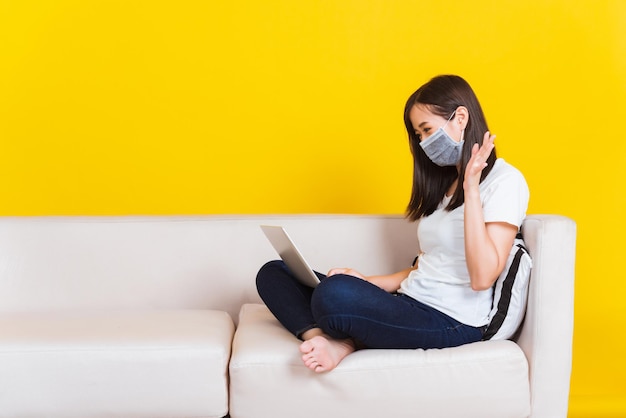 Portret Aziaat van mooie jonge vrouw zittend op de bank met medisch gezichtsmasker beschermend ze werkt vanuit huis met laptopcomputer tijdens Coronavirus studio-opname geïsoleerd op gele achtergrond