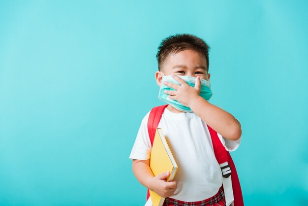 Portret aziaat weinig beschermend gezichtsmasker van de de kleuterschoolslijtage van de kindjongen en het boek van de schooltasgreep alvorens naar school te gaan