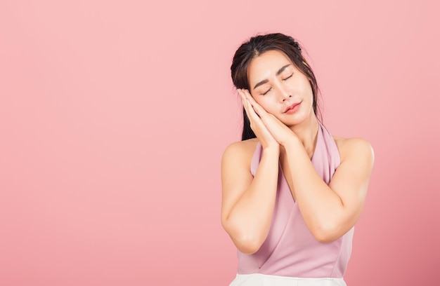 Portret Aziatische mooie jonge vrouw deed alsof emoties slapen vermoeide ogen gesloten dromen met handen samen in de buurt van gezicht, studio opname op roze achtergrond, met kopie ruimte, slapeloosheid concept