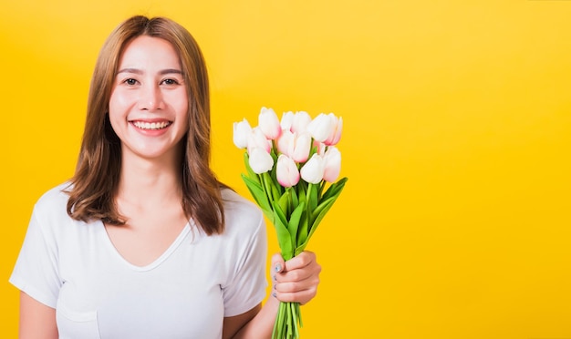 Portret Aziatische Thaise mooie gelukkige jonge vrouw glimlachend, schreeuwend opgewonden bloemen tulpen boeket in handen houden en kijken naar camera, studio opname geïsoleerd op gele achtergrond, met kopie ruimte