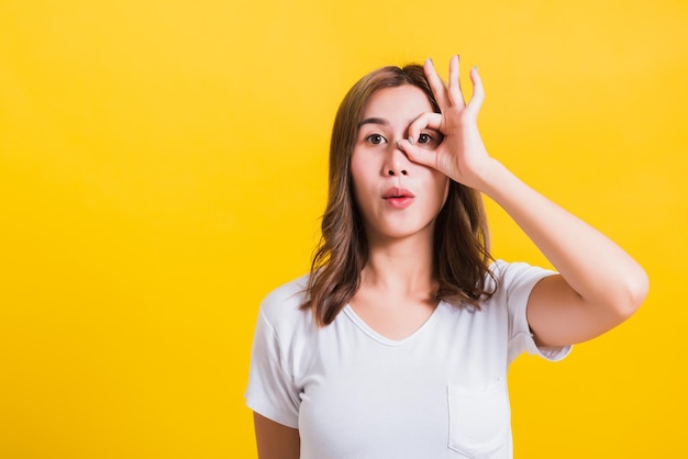 Portret Aziatische Thaise mooie jonge vrouw staande, ze maakte vinger OK symbool teken om het eens te worden in de buurt van twee ogen en kijkend naar de camera, foto schieten in de studio op gele achtergrond, er was kopieerruimte