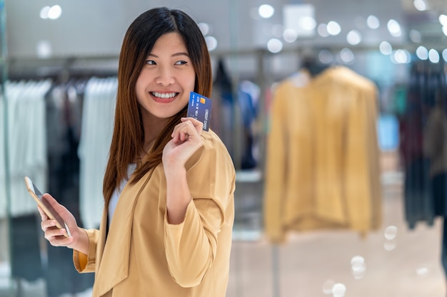 Portret Aziatische vrouw met creditcard met slimme mobiele telefoon voor online winkelen in warenhuis over de kleding winkel winkel muur, technologie geld portemonnee en online betaling concept