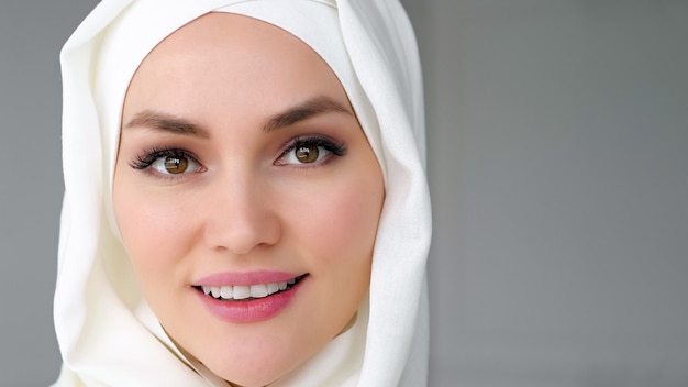 Portret close-up gezicht van mooie moslimvrouw die witte hijab draagt, kijkt naar de camera en glimlacht op een witte achtergrond, kopieer ruimte