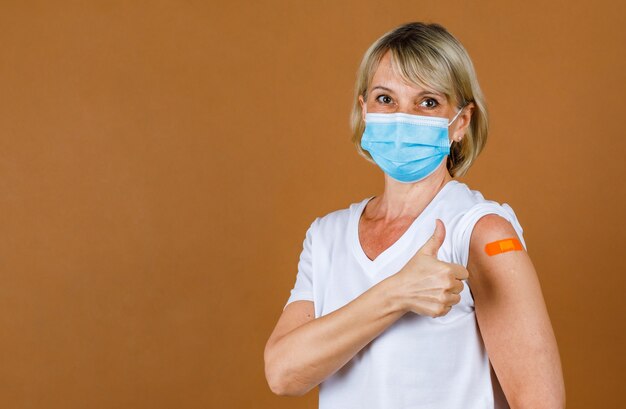 Portret Close-up studio-opname van blanke senior blonde vrouwelijke patiënt draagt gezichtsmasker, kijk naar de camera, laat duim omhoog kijken naar oranje gipsverband op haar schouder na ontvangst van Coronavirus-vaccinatie.