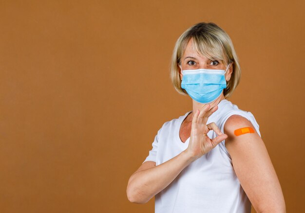 Portret Close-up studio-opname van blanke senior blonde vrouwelijke patiënt draagt gezichtsmasker, kijk naar de camera, toon handteken oke en oranje gipsverband op haar schouder na ontvangst van het Coronavirus-vaccin.