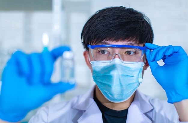 Portret close-up studio shot van Aziatische jonge professionele ervaren mannelijke wetenschapper in witte laboratoriumjas met gezichtsmasker en veiligheidsbril kijkend naar het vasthouden van een glazen injectieflacon op een wazige voorgrond.