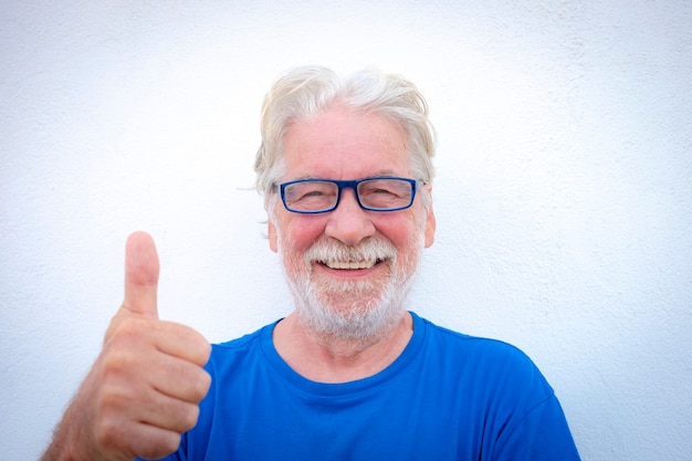 Portret close-up van een lachende senior man met witte baard op witte achtergrond met bril en tshirt in blauwe kleur. Positieve gepensioneerde persoon met duim omhoog