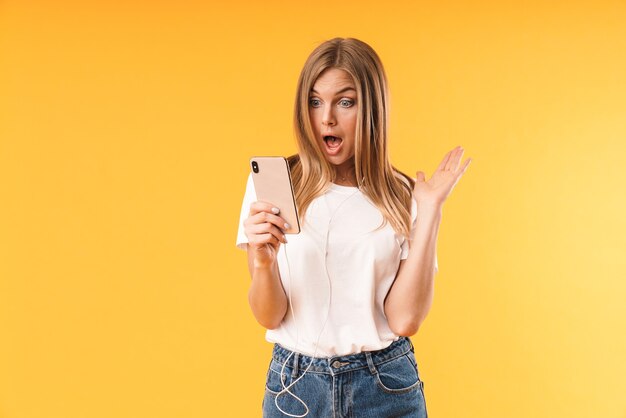 Portret close-up van opgewonden blonde vrouw die verwondering uitdrukt op smartphone tijdens het gebruik van oortelefoons geïsoleerd over gele muur