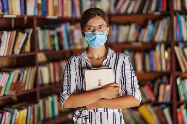 Portret dat van jong aantrekkelijk universiteitsmeisje zich in bibliotheek met gezichtsmasker bevindt, een boek houdt