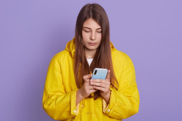 Portret die van zorgvrouw slimme telefoon in handen houden die slecht nieuws hebben, geïsoleerd stellen over lilac muur, dame in geel overhemd en lang haar heeft.