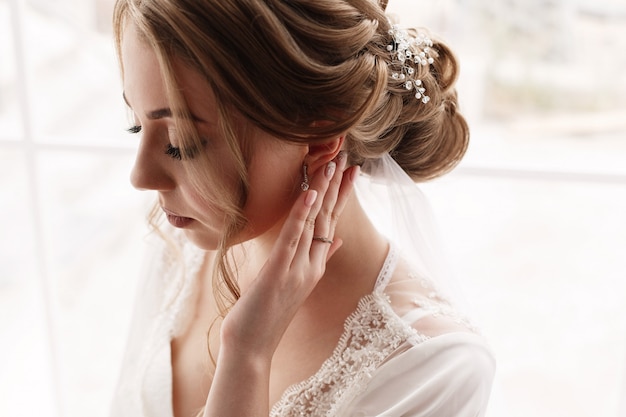 portret een mooie vrouw in witte jurk corrigeert oorbellen. tedere ochtendbruid. trouwdag. Portret van het gezicht van de schattige bruid. gezicht van jonge mooie vrouw met een mooi kapsel en make-up