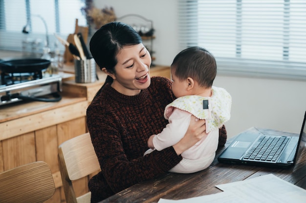 portret gelukkige aziatische freelancer moeder kijkt en speelt met haar babymeisje terwijl ze overdag thuis op de computer aan de eettafel werkt