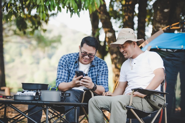 Portret gelukkige Aziatische man vrienden zittend op een stoel in het kamp met praten en kijken naar smartphone Koken set voorgrond Buiten koken reizen camping lifestyle concept