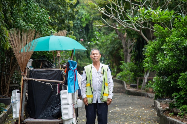 Portret gelukkige oude Aziatische man straatveger die naast een oude vuilniswagen staat voordat hij gaat werken met de natuurachtergrond