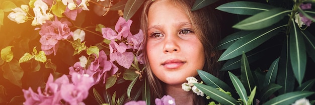 Portret gezicht van openhartig mooi klein kind meisje van acht jaar oud op achtergrond van groene planten en roze bloemen tijdens een zomervakantie reizen gen z geestelijke gezondheid concept banner flare