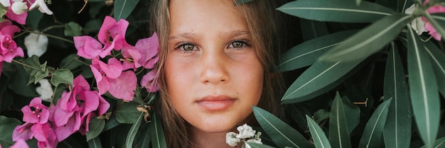 Portret gezicht van openhartig mooi klein meisje van acht jaar oud met bruine ogen op achtergrond van groene planten en roze bloemen tijdens een zomervakantie reizen gen z geestelijke gezondheid concept banner