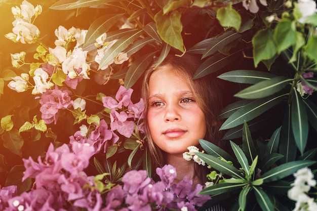 Portret gezicht van openhartig mooi klein meisje van acht jaar oud met bruine ogen op achtergrond van groene planten en roze bloemen tijdens een zomervakantie reizen gen z geestelijke gezondheid concept flare