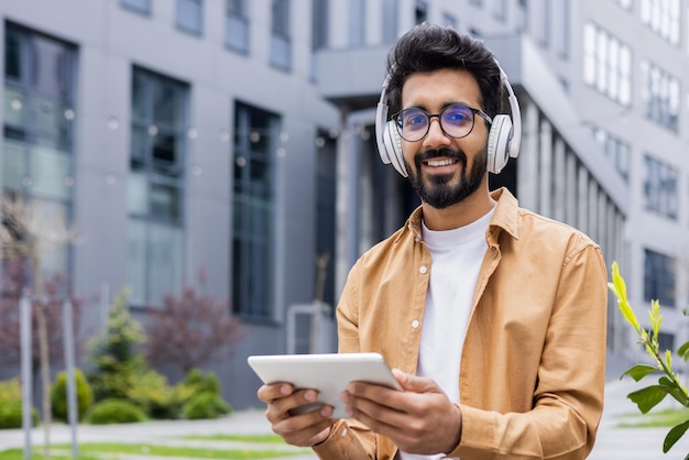 Portret jonge Indiase man met koptelefoon en tablet kijken naar online video zittend op een bankje in de buurt