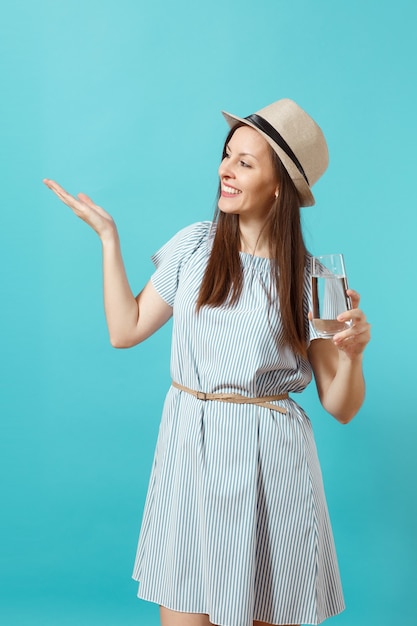 Portret jonge mooie vrouw in blauwe jurk, hoed met glas helder vers zuiver water geïsoleerd op blauwe achtergrond. Gezonde levensstijl, mensen oprechte emoties concept. Wijzende hand op kopie ruimte.